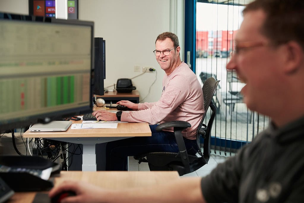 Medewerker werkt aan computer in kantoor met andere collega op de achtergrond.