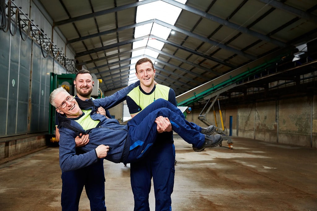 Productieleider Richard met team in de werkplaats, lachend en samenwerkend.