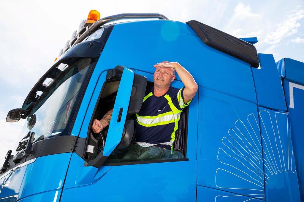 Benny, vrachtwagenchauffeur, zwaait vanuit zijn blauwe vrachtwagen.
