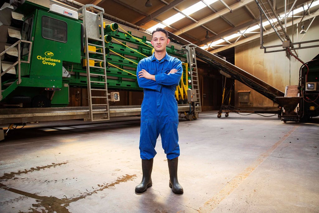 Sean, procesoperator, staat in de fabriek met industriële machines op de achtergrond.