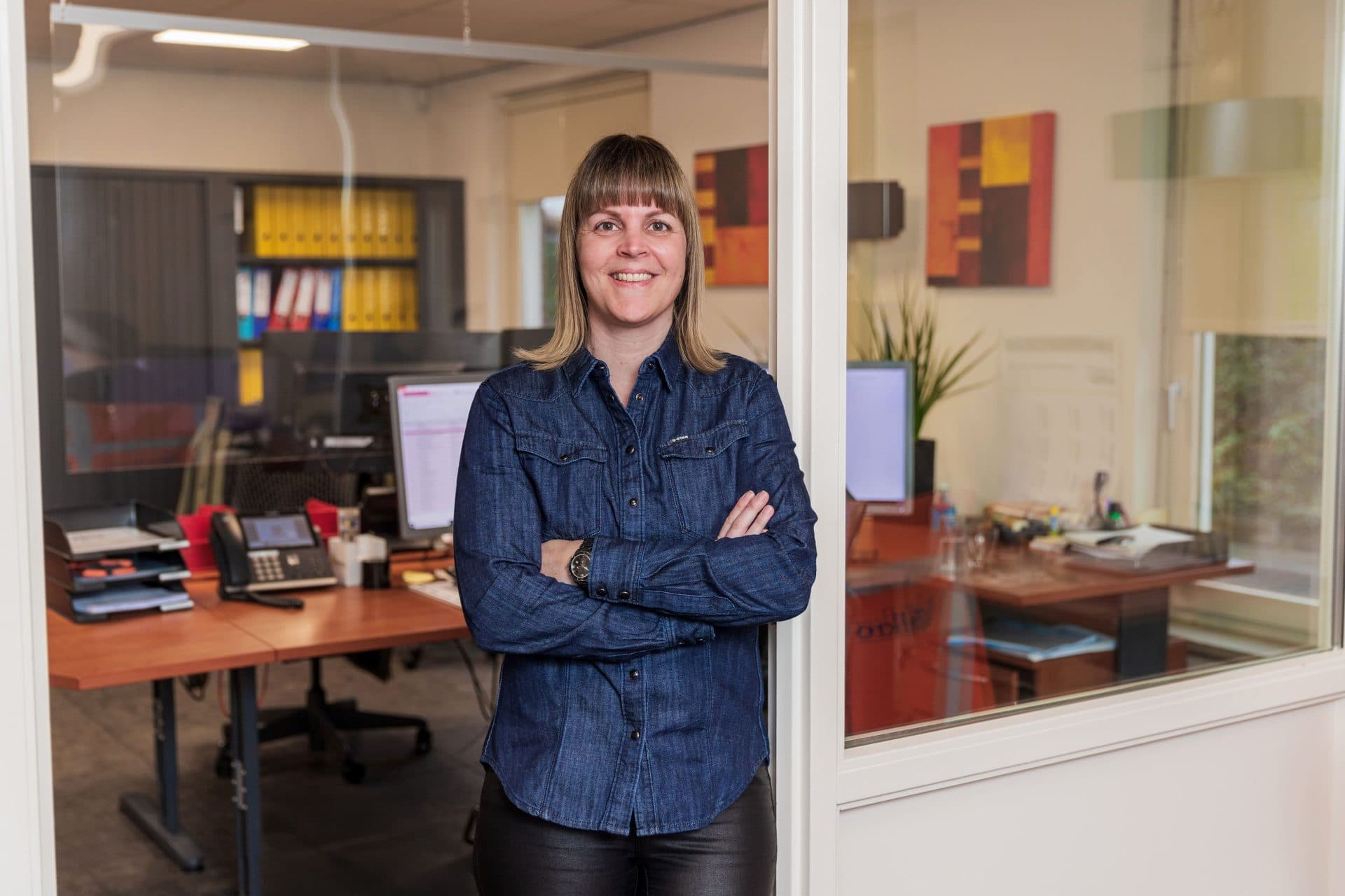 Vrouw in kantoor met armen gekruist, werkplek op achtergrond.